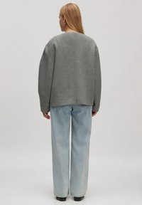 Frau mit langen blonden Haaren, die silberne Creolen, eine graue übergroße Jacke, hellblaue Schlagjeans und schwarze Schuhe trägt und mit dem Rücken zu uns steht.