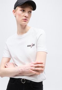 Άτομο με κοντά ξανθά μαλλιά που φοράει μαύρο καπέλο, λευκό μπλουζάκι με το κείμενο "AMOUR" και κεραυνό, μαύρο παντελόνι και ασημένια δαχτυλίδια, με σταυρωμένα τα χέρια.