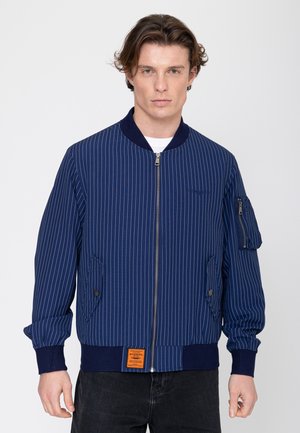 Jonge man met golvend bruin haar, die een blauwe bomberjack met krijtstreep, rits en knoopzakken draagt, staand voor een effen achtergrond.