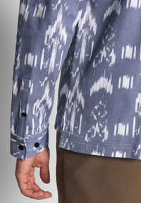 Long-sleeve shirt in blue with a white abstract pattern, featuring a buttoned cuff and textured fabric.