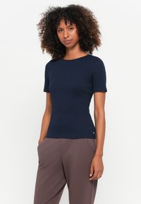 Σκούρο μπλε μπλουζάκι κοντομάνικο με στρογγυλή λαιμόκοψη, κατασκευασμένο από ένα λείο, ελαστικό ύφασμα. Αγκαλιάζει το σώμα με λεπτές λεπτομέρειες στο πλάι.