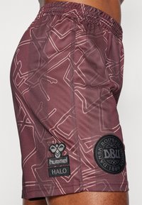 Vinröda sportshorts med ett geometriskt mönster, som har en svart "hummel HALO"-logotyp och ett runt "DBU"-märke på vänster ben.