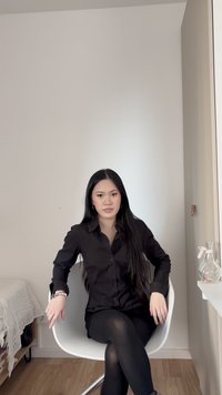 Γυναίκα με μακριά μαύρα μαλλιά, που φοράει μαύρο πουκάμισο με κουμπιά και μαύρο καλσόν, κάθεται σταυροπόδι σε μια λευκή καρέκλα σε ένα μινιμαλιστικό δωμάτιο.