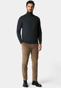 Uomo in piedi che indossa un maglione a dolcevita grigio scuro, pantaloni di velluto a coste marrone chiaro e stivali di pelle nera su uno sfondo neutro.