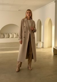 Kvinde står indendørs iført en lang beige dobbeltrækket frakke, strikketrøje, vide bukser og ankelstøvler i et minimalistisk rum med buer.