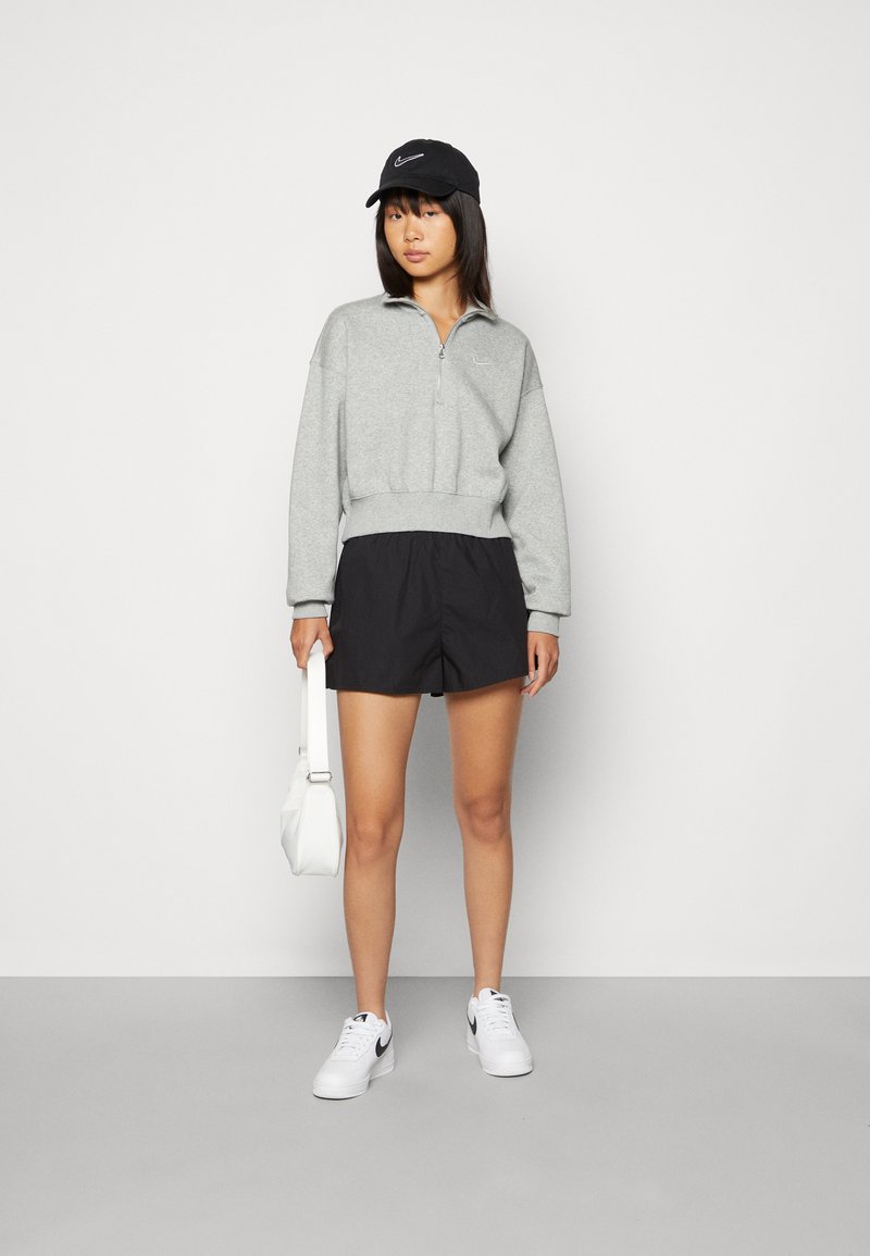 Sudadera gris con cremallera y corte holgado, combinada con pantalones cortos negros, zapatillas blancas y una gorra negra. El modelo sostiene una bolsa blanca.
