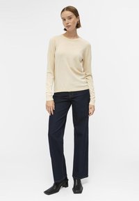 Pull en tricot beige à manches longues et col rond, associé à un jean large bleu foncé et des bottines à talons noires.