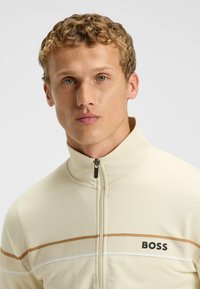 Lys beige zip-up jakke med høj krave, prydet med brune og hvide vandrette striber samt "BOSS" logoet på venstre side. Glat tekstur.