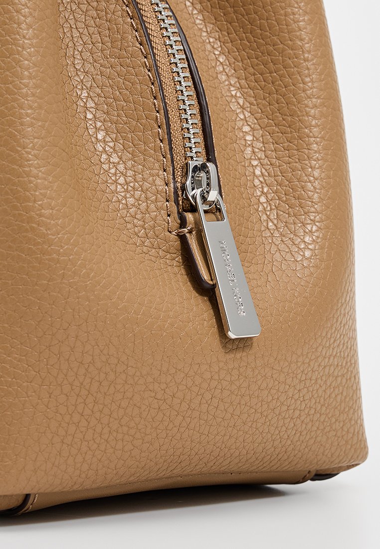 Tan läderhandväska med en strukturerad yta, har ett silverfärgat blixtlås och en rektangulär etikett med varumärkesnamnet. Närbild.