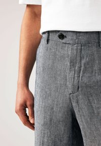 Pantalons en lin gris avec une finition texturée, dotés d'une fermeture à bouton sur le devant et de poches latérales, présentés avec un t-shirt blanc.