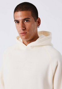 Jonge man met kort haar, gekleed in een lichtbeige hoodie met een sterlogo en dubbele ringoorbellen, kijkt recht in de camera.