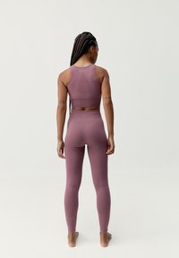 Sportinė apranga dulkių rožinės spalvos, kurią sudaro prigludęs trumpas sijonas ir aukštu liemeniu kelnes. Lygi, elastinga medžiaga su subtiliu tekstūru ir logotipo detale.