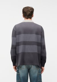 Άνδρας με κοντά καστανά μαλλιά που φοράει πόλο με μακριά μανίκια με γκρι οριζόντιες ρίγες και μπλε τζιν παντελόνια cargo, βλέπουμε από πίσω.