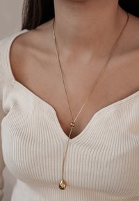 Goldener Anhänger in Tropfenform mit einer runden Perle, auf einer zarten Kette hängend, getragen über einem gerippten weißen Oberteil.