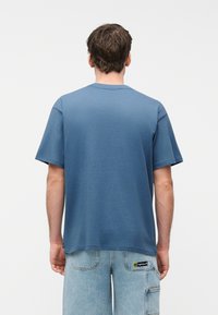 Άνδρας με καστανά κοντά μαλλιά, φοράει χαλαρό μπλε T-shirt και ανοιχτόχρωμα τζιν cargo σορτς, φαίνεται από πίσω πάνω σε απλό φόντο.