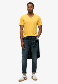 Maglietta gialla a scollo a V, jeans blu e sneakers bianche con accenti verdi. Camicia a quadri annodata in vita. Il modello è in piedi con un'espressione neutra.