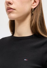 Κοντινό πλάνο ενός ατόμου που φοράει μαύρο ριμπ μπλουζάκι με ένα μικρό κόκκινο, άσπρο και μπλε λογότυπο και ένα χρυσό σκουλαρίκι κρίκο στο αριστερό αυτί.