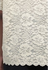 Ύφασμα από δαντέλα με χρώμα κρέμα και λουλουδάτα σχέδια, με κυματιστά άκρα πάνω σε σκούρα ρούχα, που δείχνει περίτεχνα μοτίβα λουλουδιών και φύλλων.