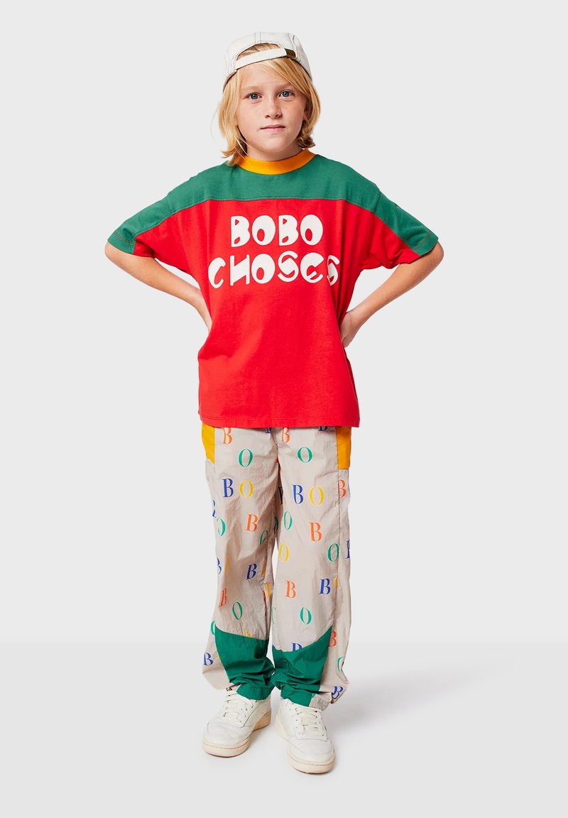 Meerkleurige outfit met een rode en groene T-shirt waarop de tekst "BOBO CHOSES" staat en patroonbeige broek. Accenten zijn oranje en groene details.