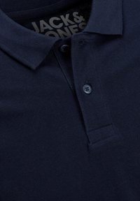 Marineblaues Poloshirt aus strukturiertem Stoff, mit einer zwei-knopfigen Knopfleiste und einem flachen Kragen. Mit einem Etikett, das im Kragen aufgedruckt ist.