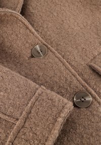 Detailweergave van een bruine, textuurvolle wollen jas met grote ronde knopen en een bijpassende textuurriem.
