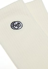 Cremfarbene, gerippte Socken mit einem schwarzen, bestickten Logo im Kreisdesign. Die Textur ist weich und glatt.