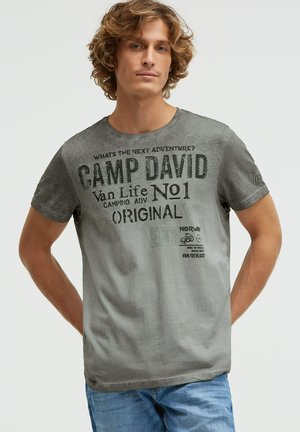Junger Mann mit lockigem Haar, der ein graues Camp David Grafik-T-Shirt und blaue Jeans trägt und mit den Händen hinter dem Rücken steht.