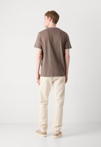 Braunes T-Shirt mit kurzen Ärmeln und lässiger Passform, kombiniert mit hellbeigen Hosen und beigefarbenen Sneakern, vor einem schlichten weißen Hintergrund.