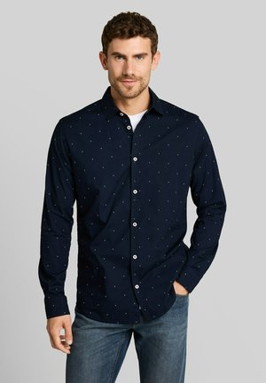 Mann mit kurzem lockigem braunem Haar, der ein dunkelblaues Hemd mit kleinen weißen Punkten und blaue Jeans trägt, steht mit einer Hand in der Hosentasche.