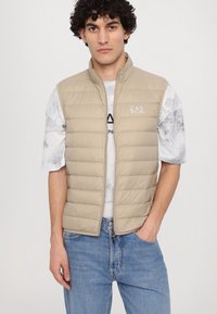 Beige, quiltet puffer vest med høy krage, glidelås og logo-detalj. Modellen har den på over en lyse mønstret skjorte og blå jeans.