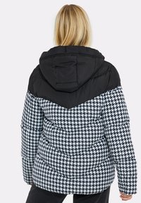 Sort-hvid houndstooth-mønstret puffers jakke med en sort, polstret hætte og skulderområde. Den har en quiltet tekstur og en tætsiddende pasform.