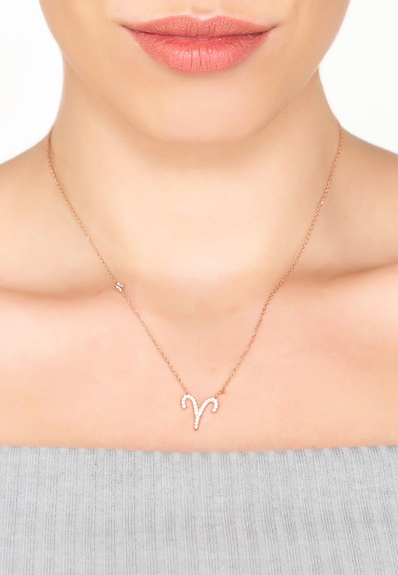 Rose gold necklace features an Aries symbol design adorned with small clear crystals, displayed on a person wearing a gray off-shoulder top.