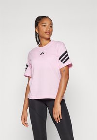 Lyserød t-shirt med korte ærmer, der har sorte tre-stribede detaljer på ærmerne og et lille sort logo på brystet. Blødt bomuldsstof.
