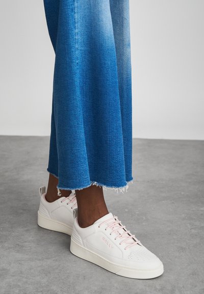 Άτομο που φοράει λευκά Bally sneakers με ροζ κορδόνια και φαρδιά μπλε τζιν παντελόνια, στέκεται σε γκρι δάπεδο.