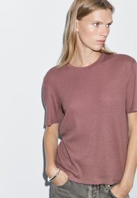 Short-sleeve, crew neck t-shirt in dusty rose color, made of soft fabric. Model hands in pockets, wearing light gray jeans and a silver bracelet.