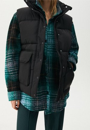 Persoon draagt een zwart donsvest over een groen, zwart en wit geruit flanellen overhemd met donkergroene broek, hand in het vestzakje.