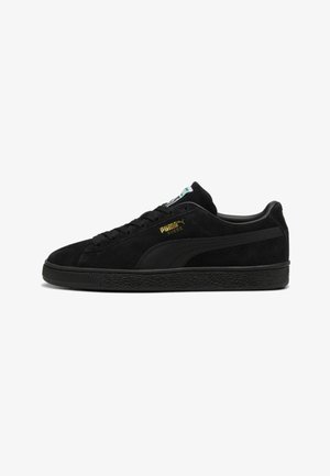 Zwarte suede sneakers met een gestructureerde zwarte rubberen zool, voorzien van een gouden logo en een zwarte streep die langs de zijkant loopt.