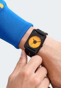 La montre rectangulaire noire présente un cadran orange, un bracelet en silicone noir et des accents orange contrastants. L'utilisateur règle l'heure à son poignet.