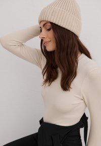 Camiseta de manga larga ribeteada en color beige con cuello redondo, combinada con un gorro de punto en beige claro. Detalles de tela negra en la cintura.