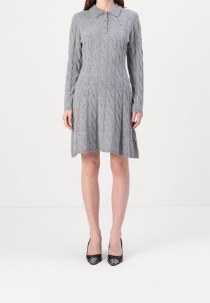 Kobieta ubrana w szarą, długorękawową, do kolan sukienkę swetrową z warkoczowym splotem i kołnierzykiem, w czarne czółenka z wąskim noskiem, stojąca na białej podłodze.
