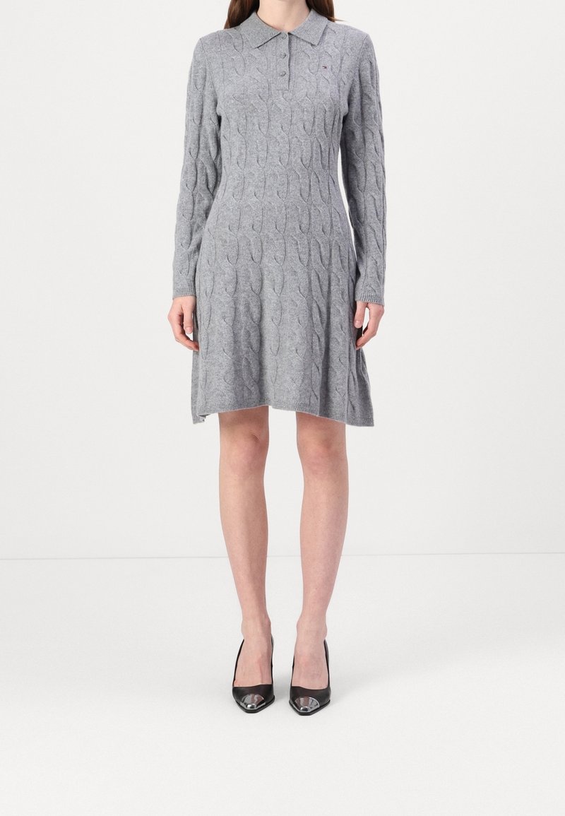 Femme portant une robe-pull grise en maille torsadée à manches longues, longueur genou, avec un col, et des escarpins noirs à bout pointu, debout sur un sol blanc.