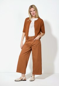Brauner, kurzer Cardigan mit gewellten Kanten, weißem Grafik-T-Shirt und weiten braunen Hosen. Abgerundet mit beigen Riemchensandalen.