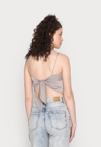 Top cinza sem costas com um detalhe de laço atado, emparelhado com jeans claros rasgados, apresentando uma etiqueta na cintura e bolsos visíveis.