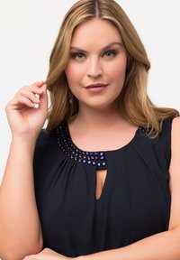 Blusa blu navy con scollatura arrotondata, caratterizzata da un'applicazione di gioielli e un'apertura a forma di chiave sul davanti. Tessuto morbido e fluido.