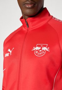 Red track jacket with a zippered front, featuring white logo and text accents. Smooth texture with contrasting details on shoulders.