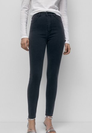 Frau trägt hoch taillierte schwarze Skinny-Jeans kombiniert mit einem weißen Langarmshirt und silbernen Riemchen-Absätzen vor schlichtem weißem Hintergrund.