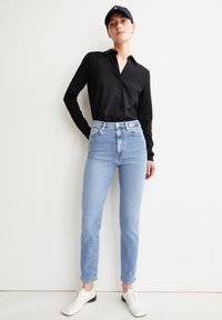 Zwarte blouse met lange mouwen, gecombineerd met lichtblauwe high-waisted jeans en witte schoenen. De blouse heeft een kraag; de jeans hebben een slim-fit ontwerp.