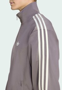 Lila träningsjacka med hög krage, vit tre-stripes design på ärmarna och en ficka med dragkedja på sidan.