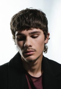 Primo piano di un giovane uomo con capelli corti e strutturati e baffi, che indossa un cappotto scuro sopra una camicia color vinaccia, su uno sfondo chiaro.