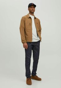 Chaqueta ligera marrón con un bolsillo en el pecho con cremallera, llevada sobre una camisa gris claro. Jeans de mezclilla oscura y zapatos marrones completan el conjunto.